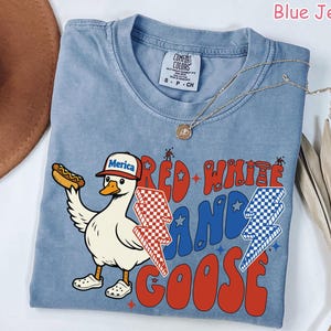 May include: Blue jean colored t-shirt with a graphic of a goose wearing a "Merica" hat and holding a hot dog. The shirt has the words "Red, White, and Goose" in red and blue. A gold necklace is draped across the shirt.