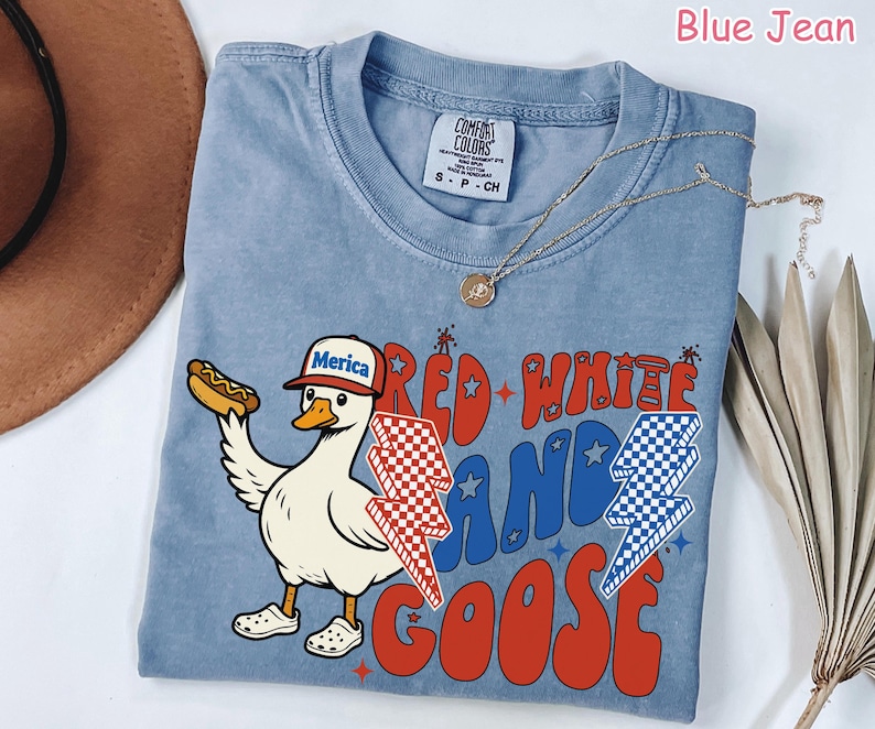 May include: Blue jean colored t-shirt with a graphic of a goose wearing a "Merica" hat and holding a hot dog. The shirt has the words "Red, White, and Goose" in red and blue. A gold necklace is draped across the shirt.