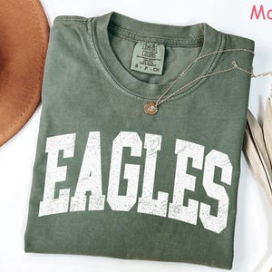 May include: A moss green t-shirt with the word "EAGLES" printed in large, distressed white letters. The shirt has a crew neck and short sleeves. A gold necklace with a pendant is draped on the shirt. The shirt is folded on a white surface.