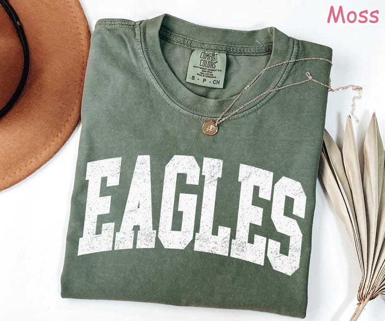 May include: A moss green t-shirt with the word "EAGLES" printed in large, distressed white letters. The shirt has a crew neck and short sleeves. A gold necklace with a pendant is draped on the shirt. The shirt is folded on a white surface.
