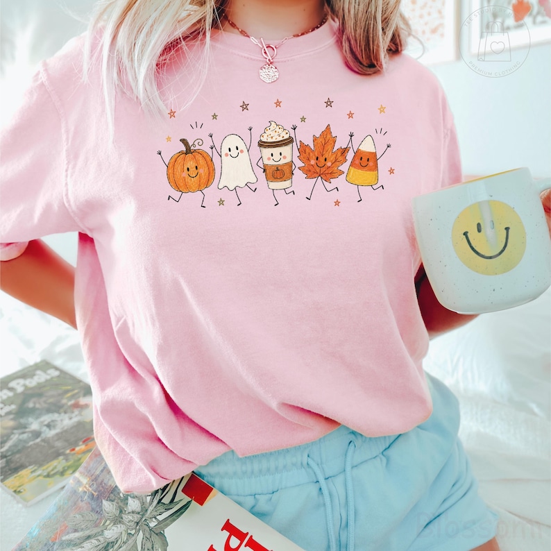 Op de afbeelding: Een lichtroze t-shirt met een grafische print van een lachende pompoen, spook, koffiebeker, esdoornblad en maissnoepje. Het shirt wordt gecombineerd met een lichtblauwe korte broek en een witte mok met een geel smiley-gezicht op de voorzijde.