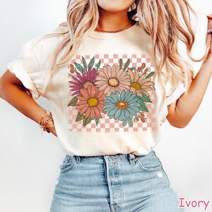 May include: Ivory-colored t-shirt featuring a colorful floral design. The graphic includes various flowers in shades of pink, orange, blue, and yellow, set against a pink and white checkered background. The shirt is made of a soft material.