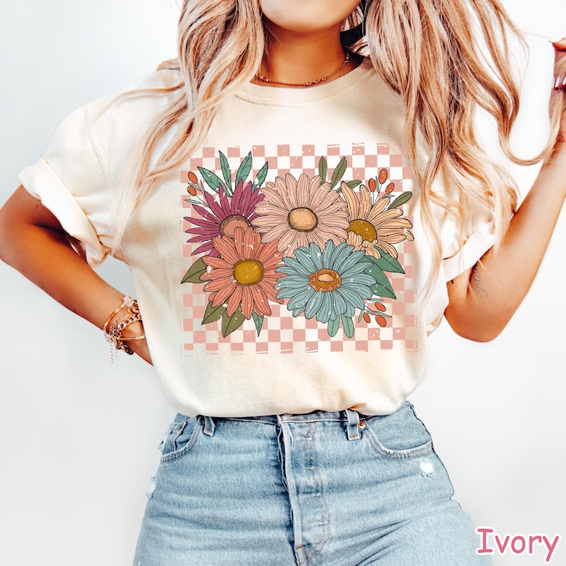 May include: Ivory-colored t-shirt featuring a colorful floral design. The graphic includes various flowers in shades of pink, orange, blue, and yellow, set against a pink and white checkered background. The shirt is made of a soft material.
