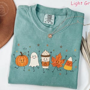 Op de afbeelding: Een lichtgroen t-shirt met een grafische print van een lachende pompoen, een spookje, een koffiebeker met slagroom, een herfstblad en een maissnoepje, allemaal voorzien van armen en benen, omringd door kleine sterretjes.