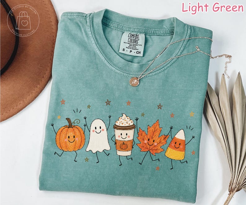 Op de afbeelding: Een lichtgroen t-shirt met een grafische print van een lachende pompoen, een spookje, een koffiebeker met slagroom, een herfstblad en een maissnoepje, allemaal voorzien van armen en benen, omringd door kleine sterretjes.