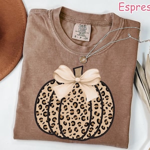 May include: Espresso-colored t-shirt featuring a pumpkin design with a leopard print pattern and a cream-colored bow. The shirt has a crew neck and short sleeves. A silver necklace with a pendant is also visible.