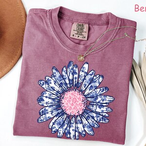 May include: Berry-colored t-shirt with a large floral design. The flower has blue and white patterned petals and a pink center. A gold necklace rests on the shirt. The shirt has a Comfort Colors tag.