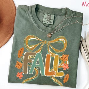 May include: A moss green t-shirt with a large graphic that says "FALL" in colorful letters. The design includes a bow and autumn leaves in shades of orange, brown, and green. A gold necklace is draped across the shirt.