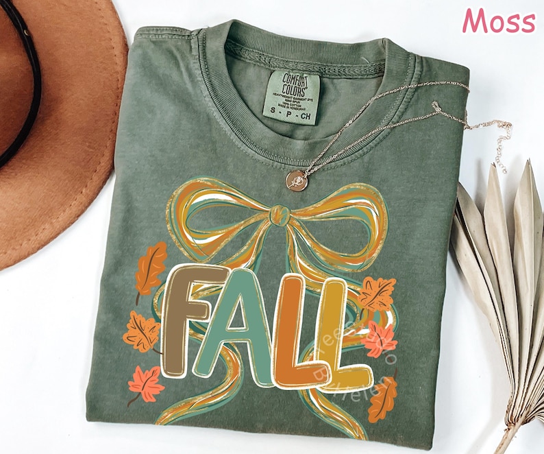 May include: A moss green t-shirt with a large graphic that says "FALL" in colorful letters. The design includes a bow and autumn leaves in shades of orange, brown, and green. A gold necklace is draped across the shirt.