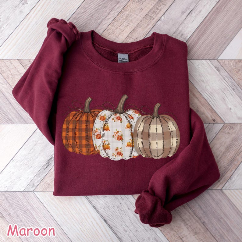 Puede incluir: Sudadera granate con tres dise&ntilde;os de calabazas: cuadros naranjas, floral y cuadros beige. Las calabazas tienen tallos verdes. La palabra "Maroon" est&aacute; impresa en la parte inferior de la imagen.