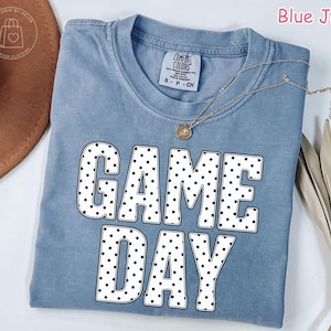Puede incluir: Una camiseta color azul jean con cuello redondo y mangas cortas. El frente presenta el texto GAME DAY en letras grandes, negritas y blancas con un patr&oacute;n de puntos negros. Un collar fino de tono dorado descansa sobre el cuello de la prenda.