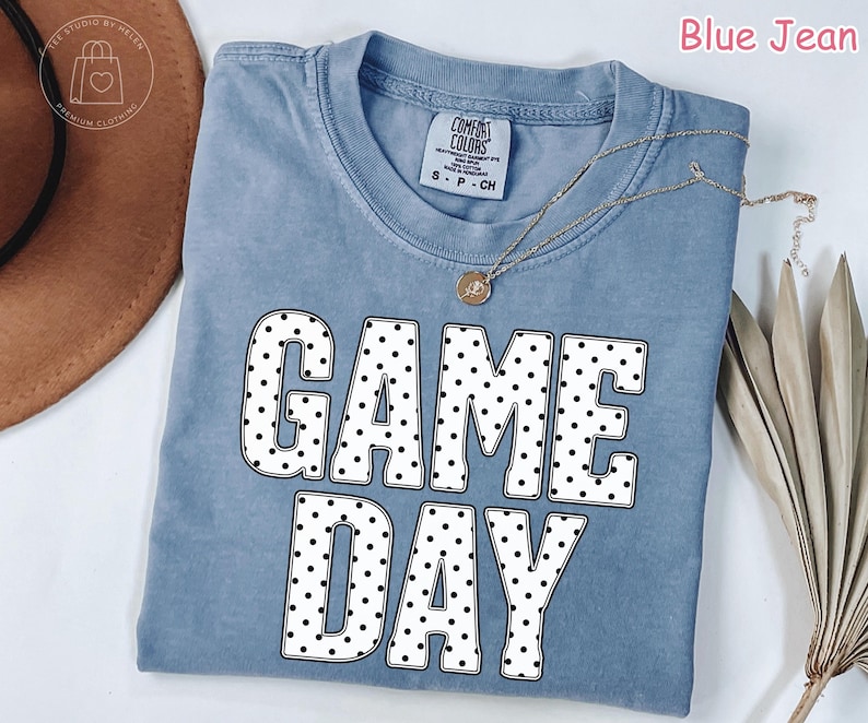 Puede incluir: Una camiseta color azul jean con cuello redondo y mangas cortas. El frente presenta el texto GAME DAY en letras grandes, negritas y blancas con un patr&oacute;n de puntos negros. Un collar fino de tono dorado descansa sobre el cuello de la prenda.