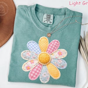 May include: Light green t-shirt with a large, colorful daisy applique. The daisy features petals in various patterns and colors, including pink gingham, blue gingham, and floral prints. A silver necklace with a small pendant rests on the shirt.
