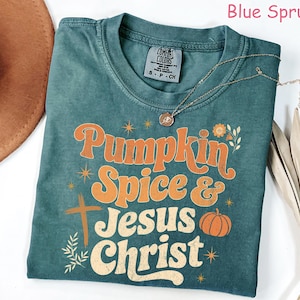 Op de afbeelding: Blauwgroen t-shirt met de woorden "Pumpkin Spice & Jesus Christ" in oranje en wit. Het shirt heeft een kruis en een pompoenafbeelding. Een gouden ketting en een bruine hoed zijn ook zichtbaar.