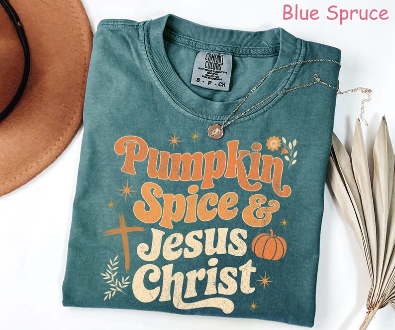 Op de afbeelding: Blauwgroen t-shirt met de woorden "Pumpkin Spice & Jesus Christ" in oranje en wit. Het shirt heeft een kruis en een pompoenafbeelding. Een gouden ketting en een bruine hoed zijn ook zichtbaar.