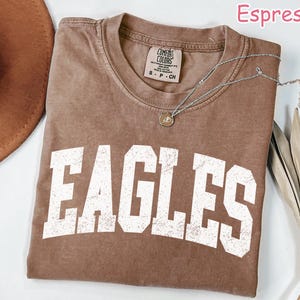 May include: Espresso-coloured t-shirt featuring the word "EAGLES" in large, distressed white letters. The shirt has a crew neck and a Comfort Colors tag. A silver necklace with a pendant is draped across the shirt.