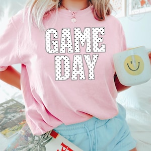 Puede incluir: Una camiseta de manga corta de color rosa claro con el texto GAME DAY en letras blancas grandes y negritas con un patr&oacute;n de puntos negros. La camiseta se combina con pantalones cortos azul claro y una taza blanca con un dise&ntilde;o de cara sonriente amarilla.