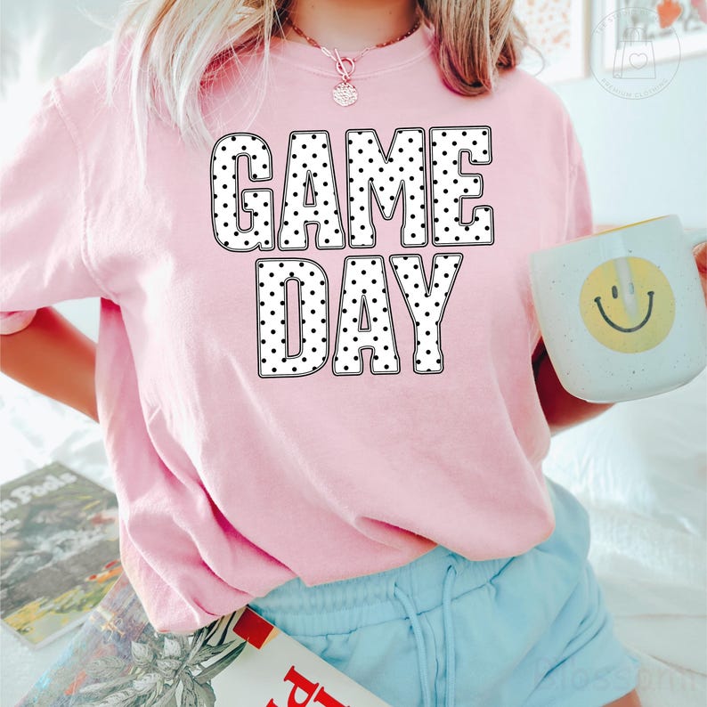 Puede incluir: Una camiseta de manga corta de color rosa claro con el texto GAME DAY en letras blancas grandes y negritas con un patr&oacute;n de puntos negros. La camiseta se combina con pantalones cortos azul claro y una taza blanca con un dise&ntilde;o de cara sonriente amarilla.