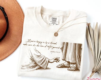 May include: A beige t-shirt with a brown illustration of a hand reaching out to a foot. The text on the shirt reads "If you're hanging on by a thread, make sure it's the hem of His garment. Matthew 12:20-27".
