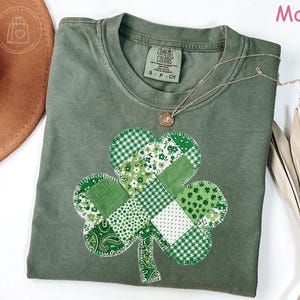 May include: A moss green t-shirt with a patchwork shamrock design. The shamrock is made of various green patterned fabrics. A gold necklace with a pendant is draped on the shirt. The shirt has a Comfort Colors tag.