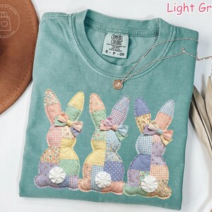 K&ouml;nnte beinhalten: Hellgr&uuml;nes Comfort Colors T-Shirt mit drei aufgen&auml;hten Patchwork-Hasen. Jeder Hase hat eine Schleife und einen wei&szlig;en Blumenschwanz. Eine silberne Halskette mit Anh&auml;nger befindet sich auf dem Shirt.
