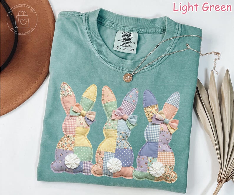 K&ouml;nnte beinhalten: Hellgr&uuml;nes Comfort Colors T-Shirt mit drei aufgen&auml;hten Patchwork-Hasen. Jeder Hase hat eine Schleife und einen wei&szlig;en Blumenschwanz. Eine silberne Halskette mit Anh&auml;nger befindet sich auf dem Shirt.