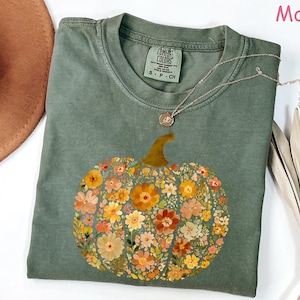 May include: A moss green t-shirt featuring a floral pumpkin design. The pumpkin is composed of various orange, yellow, and pink flowers. A silver necklace with a pendant rests on the shirt. The shirt is a Comfort Colors brand.