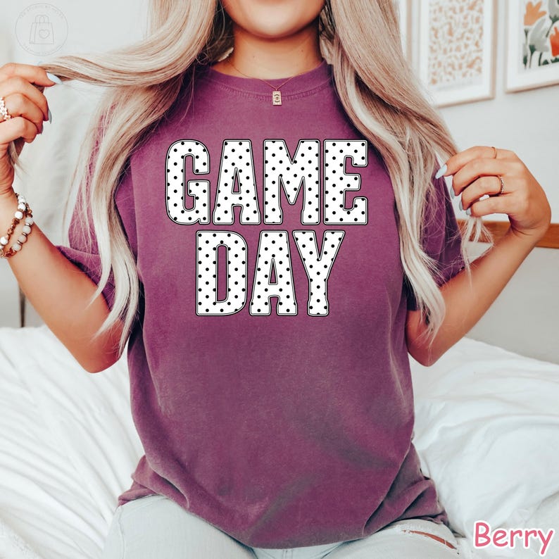 Puede incluir: Una camiseta de manga corta color baya con el texto GAME DAY impreso en una fuente blanca llamativa con un patr&oacute;n de puntos negros. La prenda tiene cuello redondo y un corte holgado, con la palabra Berry escrita en la esquina inferior derecha.