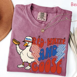 May include: Berry-colored t-shirt featuring a cartoon goose wearing a "Merica" hat and holding a hot dog. The shirt has the text "Red White and Goose" in red and blue, with lightning bolt graphics. A gold necklace is draped on the shirt.