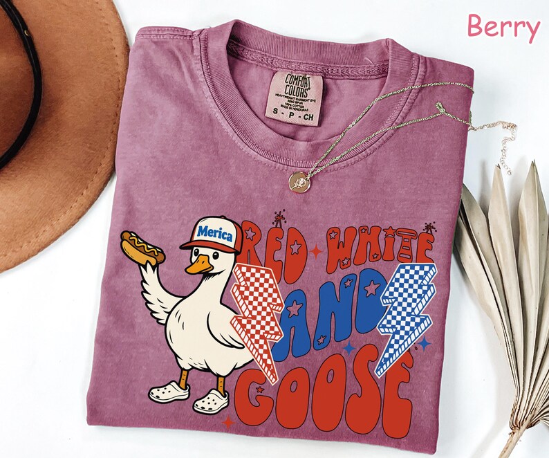 May include: Berry-colored t-shirt featuring a cartoon goose wearing a "Merica" hat and holding a hot dog. The shirt has the text "Red White and Goose" in red and blue, with lightning bolt graphics. A gold necklace is draped on the shirt.