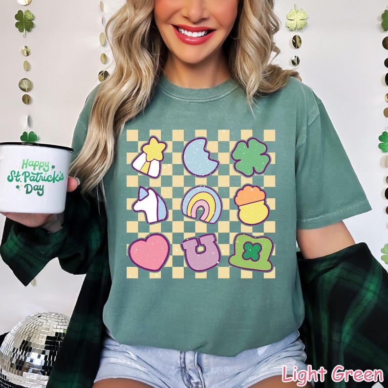 May include: A light green t-shirt with a checkered pattern and colorful cartoon images of a shamrock, a horseshoe, a rainbow, a pot of gold, a unicorn, a star, a crescent moon, and a leprechaun hat. The shirt has the text "Happy St. Patrick's Day" on it.