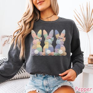 May include: Dark gray sweatshirt featuring three patchwork bunny designs with pastel colors and bows. The bunnies have a quilted appearance, with each one showcasing a unique pattern. The word "Pepper" is visible in the bottom right corner.