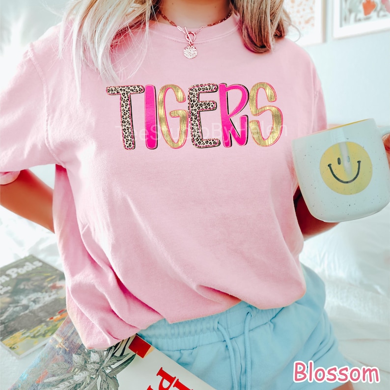 Peut inclure: Un sweat-shirt rose clair avec le mot "TIGERS" dans un design audacieux et superpos&eacute;. Les lettres combinent un imprim&eacute; l&eacute;opard, rose vif et or. Une tasse blanche avec un smiley jaune est tenue dans la main.