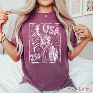 Puede incluir: Una camiseta de manga corta color baya con un gr&aacute;fico de l&iacute;neas blancas de la Estatua de la Libertad sosteniendo una antorcha y una bandera estadounidense. El dise&ntilde;o incluye el texto USA y 250, rodeado de estrellas dentro de un borde rectangular.