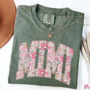 K&ouml;nnte beinhalten: Moosgr&uuml;nes T-Shirt mit dem Wort "MAMA" in einem floralen Design. Die Buchstaben sind mit rosa, wei&szlig;en und orangefarbenen Blumen gef&uuml;llt. Eine goldene Halskette mit Anh&auml;nger befindet sich auf dem Shirt.