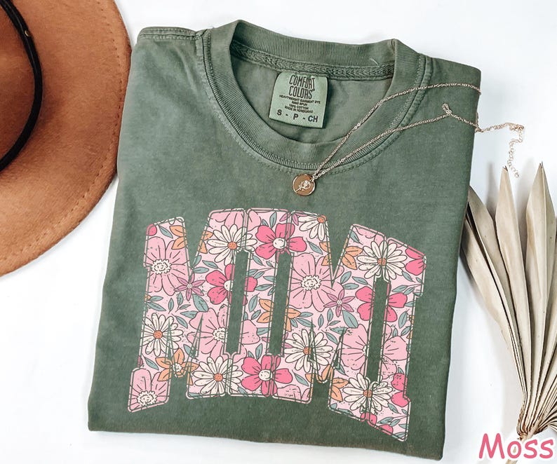 K&ouml;nnte beinhalten: Moosgr&uuml;nes T-Shirt mit dem Wort "MAMA" in einem floralen Design. Die Buchstaben sind mit rosa, wei&szlig;en und orangefarbenen Blumen gef&uuml;llt. Eine goldene Halskette mit Anh&auml;nger befindet sich auf dem Shirt.