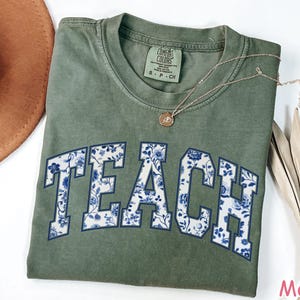 May include: A green t-shirt with the word "TEACH" printed in white with a blue floral pattern. The shirt is made of a soft, comfortable material.