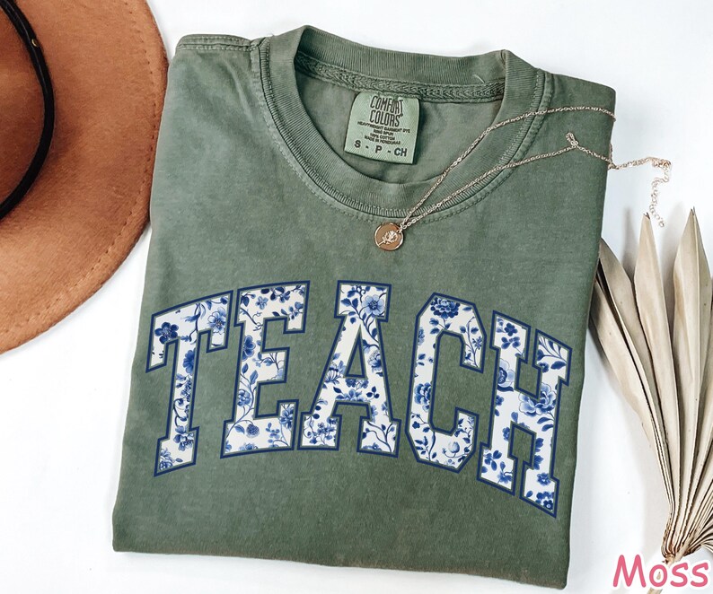 May include: A green t-shirt with the word "TEACH" printed in white with a blue floral pattern. The shirt is made of a soft, comfortable material.