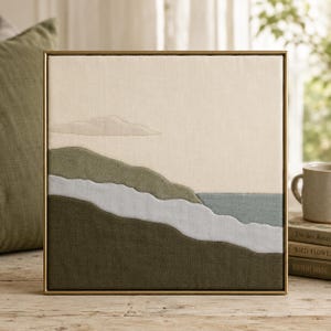 May include: Square framed wall art featuring a landscape scene. The artwork depicts a coastal view with layered fabric in shades of green, blue, and cream. A gold-colored frame surrounds the artwork, which is displayed on a wooden surface.