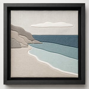 May include: A framed textile art piece depicting a coastal scene. The artwork features a beach with light blue and beige tones, a dark blue ocean, and a light gray sky with a white cloud. The frame is black.