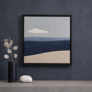 May include: Textured wall art featuring a landscape design. The artwork depicts a sky blue background with a white cloud, and layered blue and beige fabric representing the sea and sand. The artwork is framed in a black square frame.