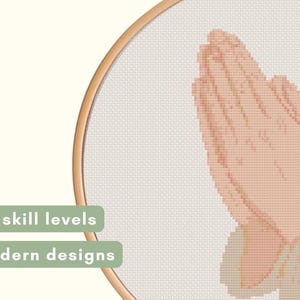May include: Cross-stitch design featuring hands clasped in prayer, rendered in a pixelated style. The design is set within a wooden embroidery hoop. Text on the left reads "All skill levels" and "Modern designs".