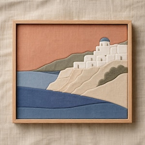 May include: A framed textile art piece depicting a coastal village scene. The artwork features white buildings with blue domes on a hillside, overlooking blue waters. The sky is a warm orange, and the frame is light wood.