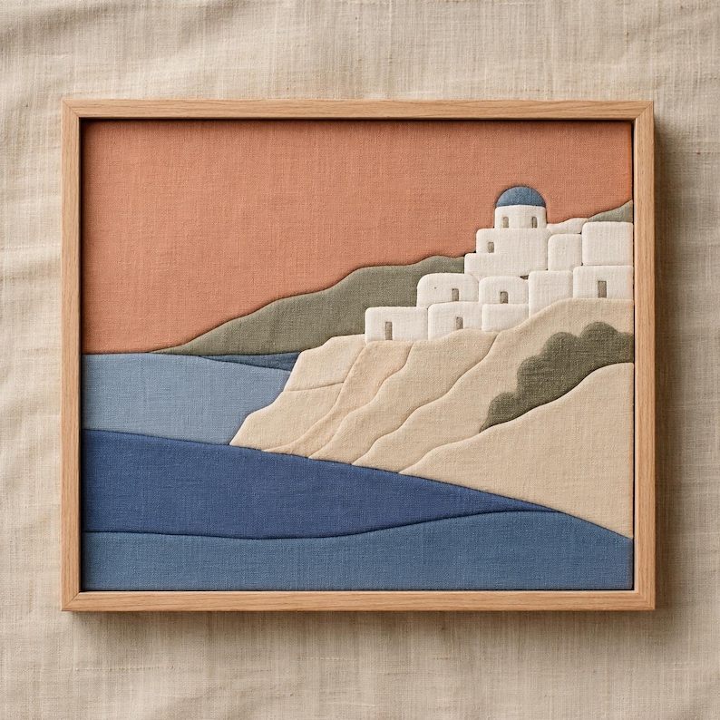 May include: A framed textile art piece depicting a coastal village scene. The artwork features white buildings with blue domes on a hillside, overlooking blue waters. The sky is a warm orange, and the frame is light wood.