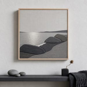 May include: A framed grayscale artwork depicting a serene coastal scene. The artwork features a textured, fabric-like appearance, with a shoreline of dark gray rocks and a calm sea reflecting the sky. The frame is a light wood color.