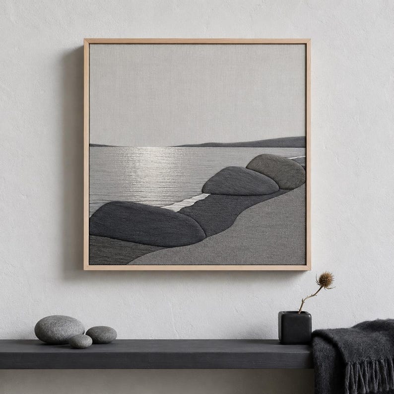May include: A framed grayscale artwork depicting a serene coastal scene. The artwork features a textured, fabric-like appearance, with a shoreline of dark gray rocks and a calm sea reflecting the sky. The frame is a light wood color.