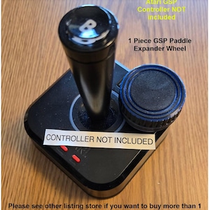 May include: Black Atari joystick with a blue and black paddle expander wheel. The text "CONTROLLER NOT INCLUDED" is on a white label attached to the joystick base. The text "Atari GSP Controller NOT included" and "1 Piece GSP Paddle Expander Wheel" is written in yellow on a wooden background.