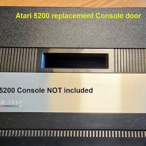 Può includere: Sportello di ricambio per console Atari 5200. Lo sportello è nero e argento con la scritta "Atari 5200 replacement Console door" e "Atari 5200 Console NOT included". Il logo Atari è visibile.