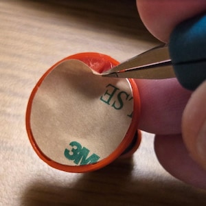 May include: An orange, circular object with a white backing and the text "3M" in green is being cut with scissors. The object has a red border and the text "SE" is partially visible. The scissors have blue handles.