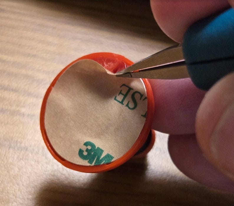 May include: An orange, circular object with a white backing and the text "3M" in green is being cut with scissors. The object has a red border and the text "SE" is partially visible. The scissors have blue handles.
